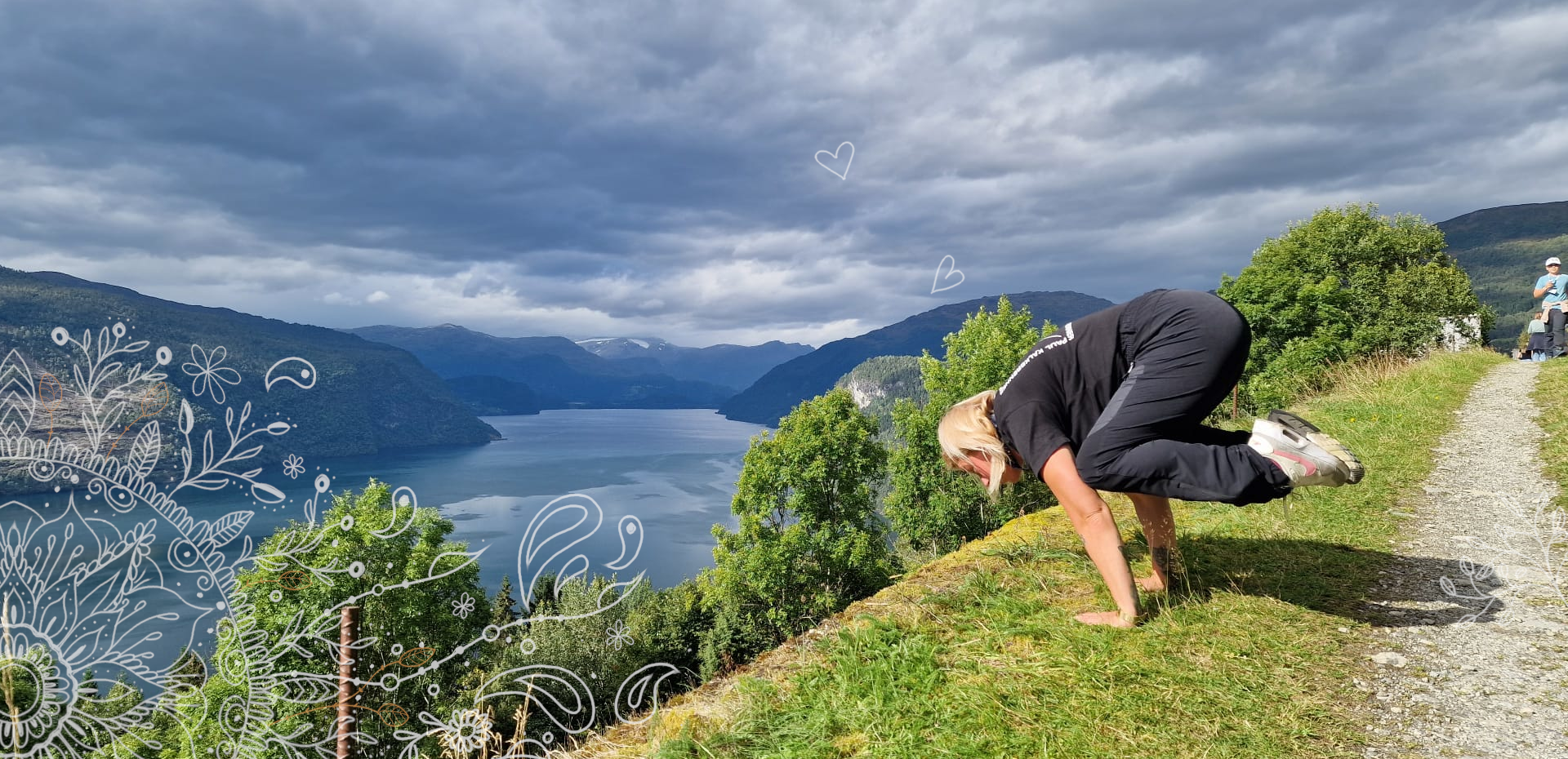 Frau in Yoga-Pose auf den Händen auf einer Wiese. Im Hintergrund ein bewölkter Himmel, Hügel, ein Fluss und Büsche.